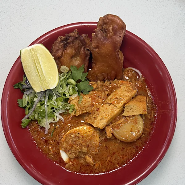 A red bowl of mohinga at Laksa with fried vegetable tempura, a lime wedge, scallions, tofu, and a half boiled egg in deep red broth