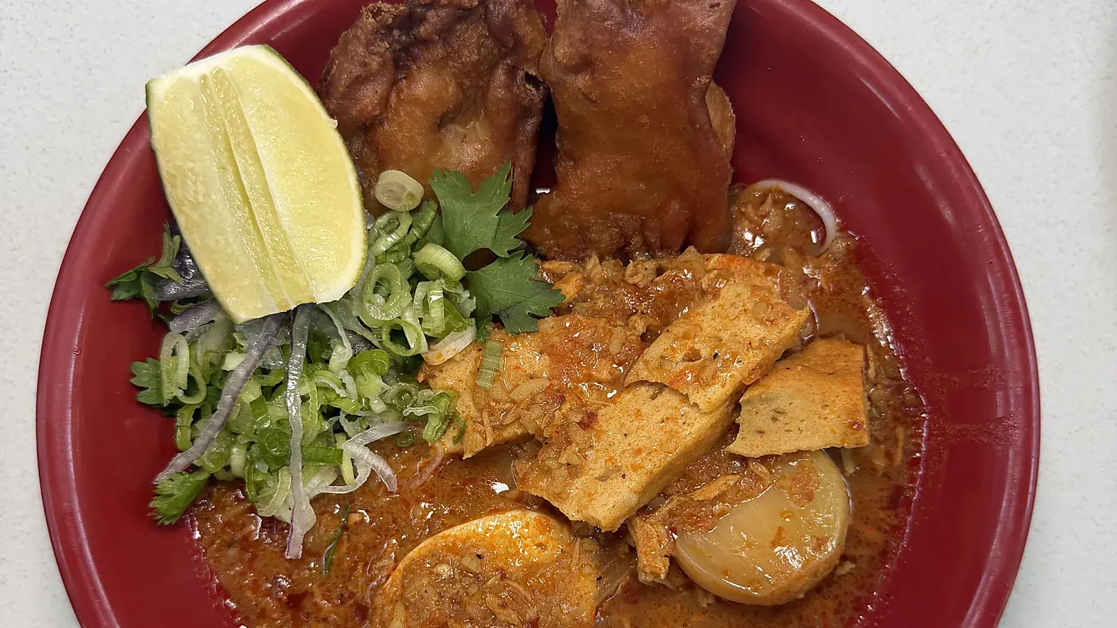A red bowl of mohinga at Laksa: rice vermicelli in a deep red broth with fried vegetable tempura, a lime wedge, sliced scallions, cilantro, slabs of tofu, and a half boiled egg