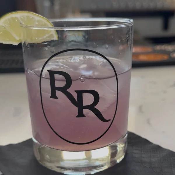 The Purple Rain Rita, a pale purple cocktail over ice in a Roch's Reprieve RR-logo rocks glass with a lime wedge on the rim