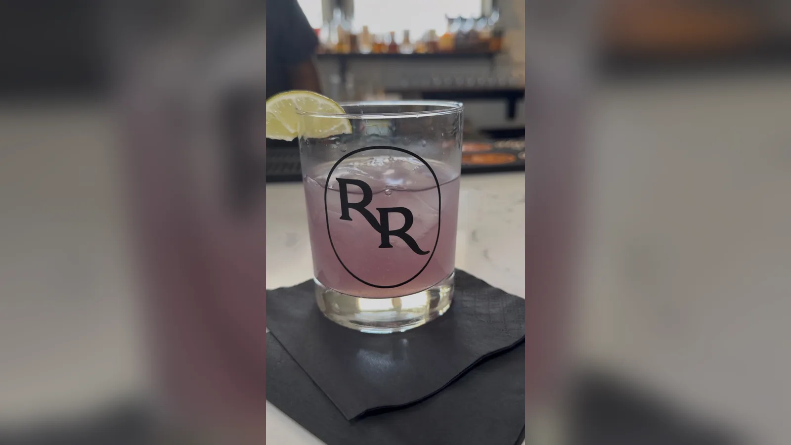 The Purple Rain Rita, a pale purple cocktail over ice in a Roch's Reprieve RR-logo rocks glass with a lime wedge on the rim, sitting on a black napkin on the bar