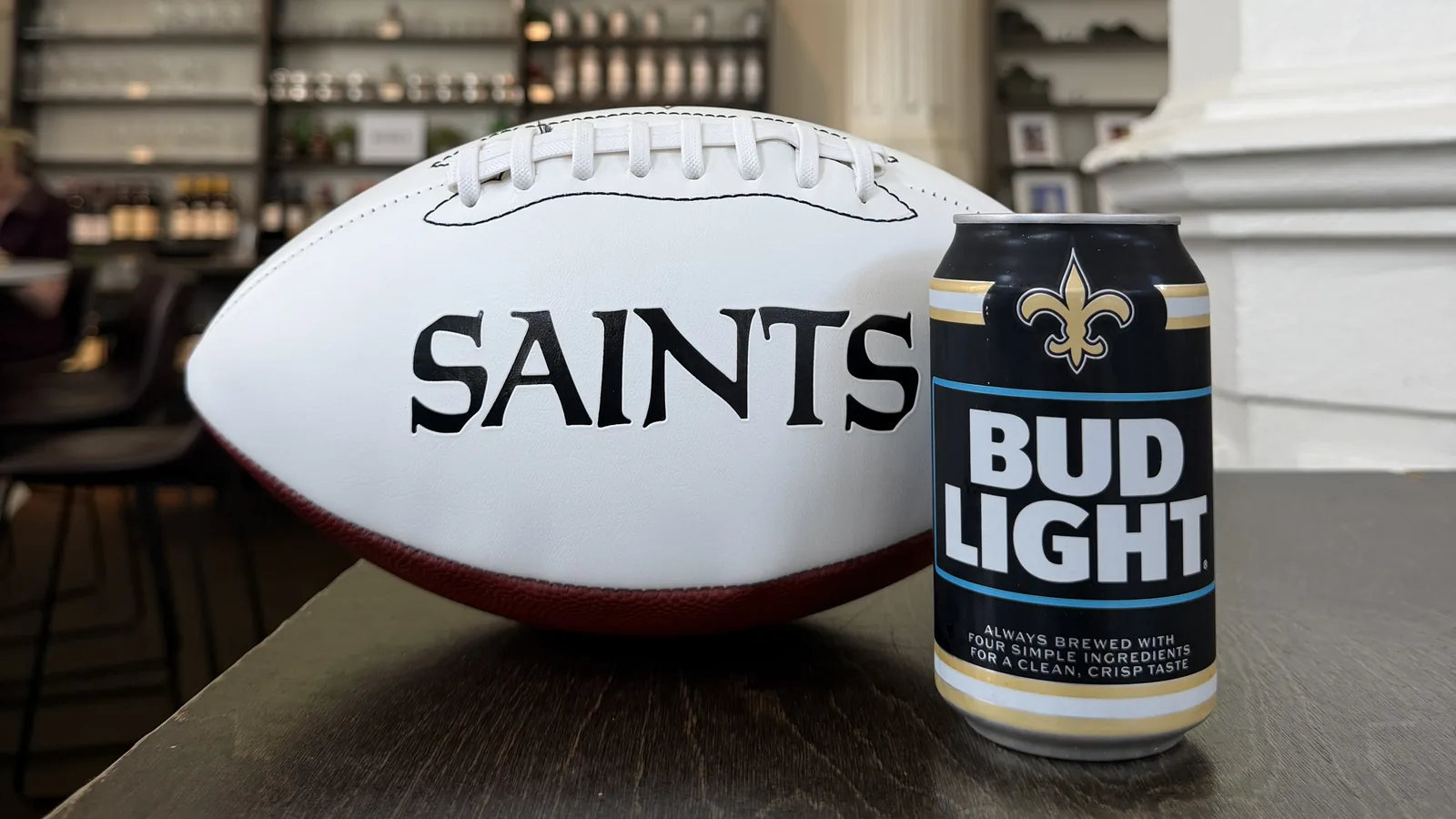 A white Saints football and a black-and-gold Saints edition Bud Light can side by side on a dark wooden table inside St. Roch Market