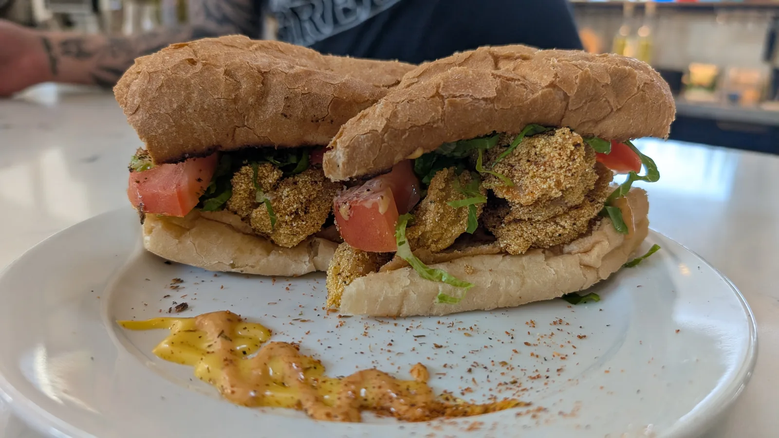 A fried oyster po-boy cut in half on a white plate at Salted Pearl Oyster Bar, cornmeal-crusted oysters with shredded lettuce and tomato on French bread, with remoulade and Creole seasoning swiped across the plate